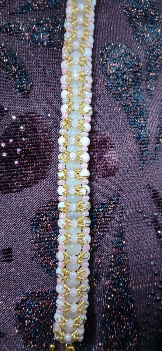 A close-up of a beaded trim or bracelet laid vertically on a textured, sparkly purple fabric background. The trim features rows of small white beads, pale pink beads, and metallic gold beads.